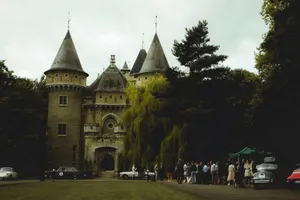 A beautiful stone castle with vintage cars and people gathered outdoors on a cloudy day. jigsaw puzzle