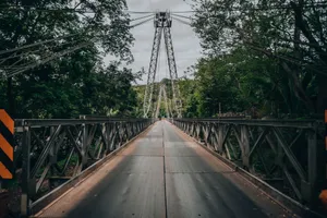 A picturesque metal suspension bridge surrounded by green trees under a cloudy sky, offering a tranquil nature escape. jigsaw puzzle