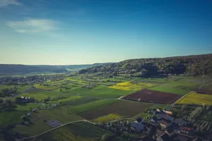 Aerial view of lush green farm fields with a distant village and clear blue sky. jigsaw puzzle