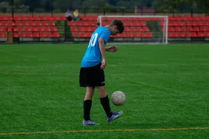 Teenage soccer player skillfully balancing a soccer ball on a grassy field with red stands. jigsaw puzzle