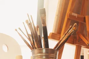 A collection of used paintbrushes in a metal container set in a warm, sunlit art studio. jigsaw puzzle