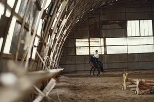 A woman rides a horse in a rustic indoor riding hall surrounded by wooden structures. jigsaw puzzle