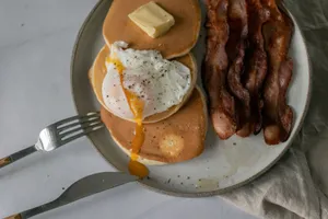 Top view of delicious pancakes with poached egg with seasoning and butter piece near sliced fried bacon on plate near fork and knife near napkin on white table jigsaw puzzle