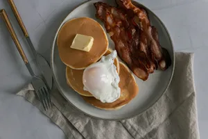 Top view of delicious pancakes with butter piece and poached egg near sliced fried bacon on plate near fork and knife near napkin on white table in bright place jigsaw puzzle