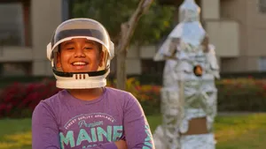 A cheerful boy wearing a space helmet stands outdoors with a homemade rocket. jigsaw puzzle